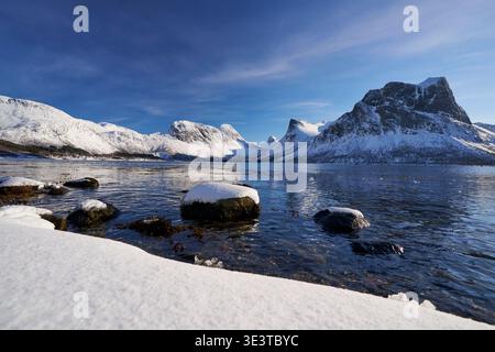 Schneebedeckte Felsen an der arktischen Fjordküste mit Bergen und klarem blauem Himmel in Norwegen, Winterlandschaft mit Reflexionen. Stockfoto