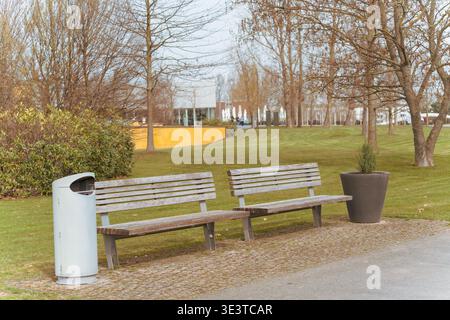 Zwei Holzbänke blicken auf einen grasbewachsenen Park. Neben ihnen steht weißer Metallmüll. Blattlose Bäume und Sträucher umgeben die Szene. Gelbes Gebäude ap Stockfoto