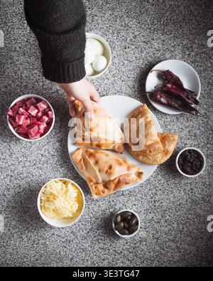 Die Hand hält Brot, umgeben von frischen Belägen und Gewürzen. Auf der Steinoberfläche befinden sich Schüsseln mit gehackten Gemüse und Oliven. Bunte Zutaten deuten auf einen Krieg hin Stockfoto