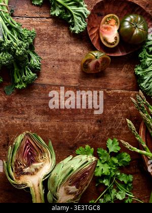 Frisches Gemüse wird auf einem rustikalen hölzernen Hintergrund platziert. Es gibt Artischocken, Tomaten, Spargel und grüne Kräuter, die zum Kochen eines veganen Gerichts bereit sind. Stockfoto