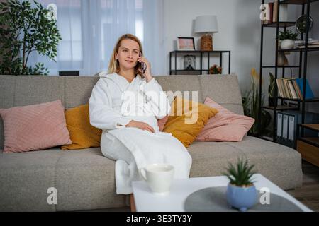 Frau, die auf dem Smartphone im Bademantel spricht und sich zu Hause auf der Couch entspannt Stockfoto