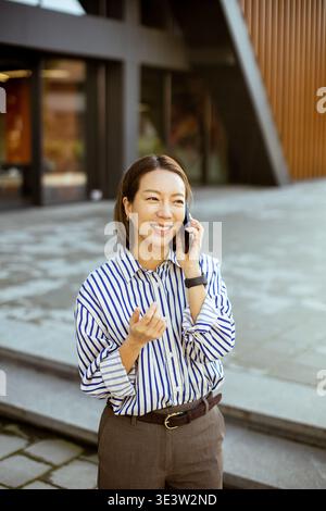 Eine Koreanerin steht vor einem modernen Gebäude. Sie lächelt, während sie am Telefon spricht. Die Anlage hat Steintreppen und eine Holzfassade. Es ist tagsüber Stockfoto