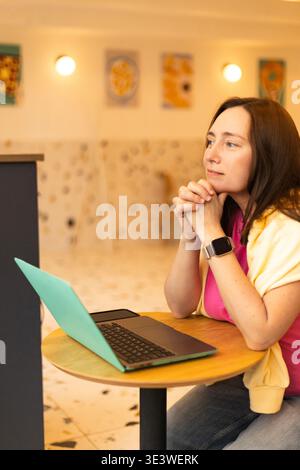 Eine junge Frau sitzt nachdenklich an ihrem Laptop und schaut aus dem Fenster. Ihr Ausdruck ist reflektierend und zeigt tiefe Gedanken oder Nachsicht in einem Casu an Stockfoto