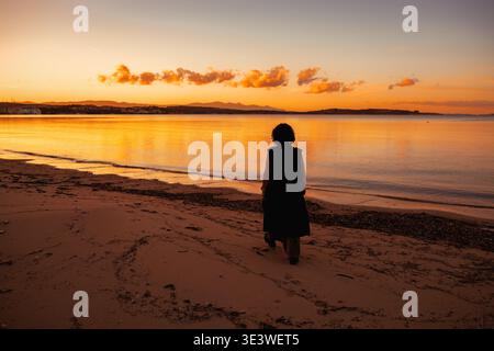 Eine Frau läuft entlang der Sandküste, die sich vor den leuchtenden Farben eines Sonnenuntergangs abhebt. Das ruhige Meer spiegelt die goldenen und orangen Töne der SK wider Stockfoto