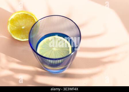 Ein lebhaftes Bild mit einem klaren blauen Glas Wasser mit einer frischen Zitronenscheibe, die im Inneren schwebt und Schatten auf eine helle Oberfläche wirft. Perfekt für Refre Stockfoto