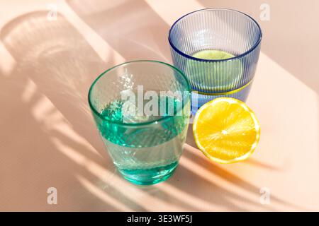 Zwei klare Gläser, eins blau und eins grün, gefüllt mit erfrischendem Wasser und garniert mit Zitronenscheiben, werfen sanfte Schatten auf eine sonnenbeschienene Oberfläche, Epi Stockfoto