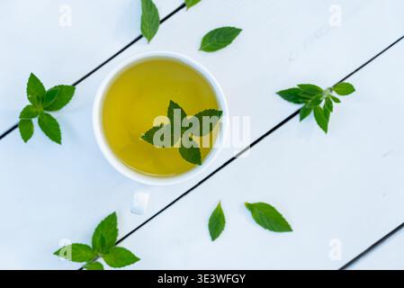 Eine Tasse frisch gebrühter, hausgemachter Minztee mit leuchtenden Minzblättern auf einer weißen Holzoberfläche bietet ein erfrischendes und aromatisches Getränk Stockfoto