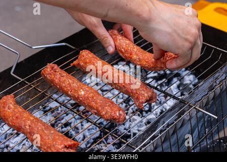 Nahaufnahme von Händen, die rohe Kofta-Kebabs auf einem Grill über heißen Kohlen platzieren. Die Szene fängt die Essenz der Outdoor-Küche und des traditionellen Middl ein Stockfoto