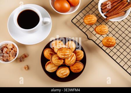 Ein Teller mit goldenen oreshki-Keksen neben einer Tasse schwarzen Kaffee, begleitet von Zutaten wie Zimtstangen, braunem Zucker und Eiern auf einer neutralen Ba Stockfoto