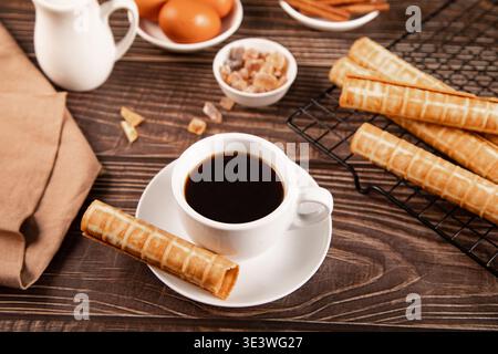 Eine dampfende Tasse schwarzen Kaffee wird mit knusprigen gerollten Waffelkeksen auf einem rustikalen Holztisch kombiniert. Zutaten wie Eier, Zucker und Zimt fügen ein h hinzu Stockfoto