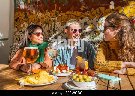Freunde genießen eine angenehme Zeit und authentische Verbindung bei Tapas und Getränken in einer gemütlichen Umgebung im Freien. Sie teilen ein Lächeln und lebhafte Gespräche Stockfoto