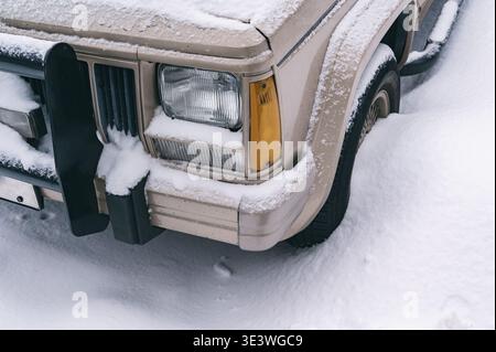 Ein Fahrzeug ist nach einem Wintersturm teilweise unter einer dicken Schneeschicht auf einer Stadtstraße vergraben. Schnee bedeckt die städtische Umgebung und wartet auf Reinigung Stockfoto