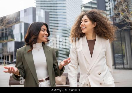 Zwei professionelle Frauen gehen und unterhalten sich selbstbewusst in einem modernen Stadtgebiet, in stilvoller Business-Kleidung. Sie scheinen Kollegen oder Freunde zu sein Stockfoto