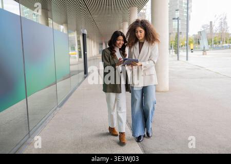 Zwei professionelle Frauen laufen zusammen vor einem modernen Bürogebäude und unterhalten sich. Sie wirken fokussiert und kooperativ, verkörpern das Team Stockfoto