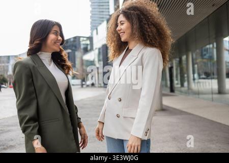 Zwei professionelle Frauen führen in einem urbanen Ambiente ein freundliches Gespräch im Freien. Beide tragen Geschäftskleidung und spiegeln Zusammenarbeit wider Stockfoto