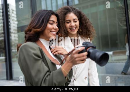 Zwei Geschäftsfrauen schauen sich gemeinsam ein Kamerafoto an, lächelnd und fesselnd. Sie stehen vor einem modernen Bürogebäude und repräsentieren die Te Stockfoto