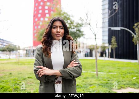 Professionelle Geschäftsfrau im grünen Blazer steht selbstbewusst in einem modernen Stadtpark. Im Hintergrund befinden sich moderne Gebäude und üppiges greene Stockfoto