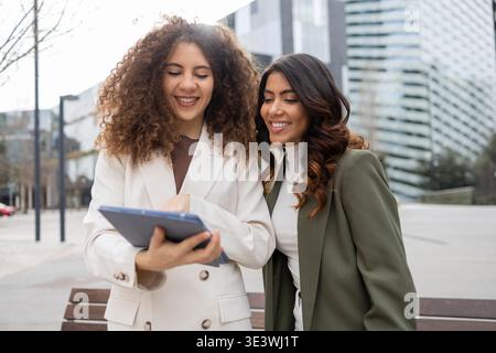 Zwei professionelle Frauen in Geschäftskleidung arbeiten mit einem digitalen Tablet im Freien zusammen und demonstrieren Teamarbeit und Freundschaft in einem urbanen Umfeld Stockfoto