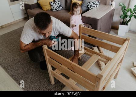Ein Mann baut geschickt mit einer Bohrmaschine ein hölzernes Babybett in einem gemütlichen Wohnzimmer zusammen, während ein neugieriges Kleinkind aufmerksam beobachtet und lernt f Stockfoto