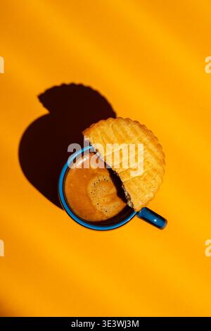 Ein Blick von oben auf eine blaue Kaffeetasse mit aufschäumendem Espresso, teilweise bedeckt von einem runden Keks, vor einer leuchtend gelben Kulisse, die einen DRAM ausstrahlt Stockfoto