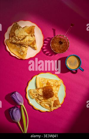 Blick von oben auf köstliche Crepes mit Fruchtmarmelade auf bunten Tellern, begleitet von einer Tasse Kaffee. Hellrosa Hintergrund mit Tulpen verstärkt das c Stockfoto