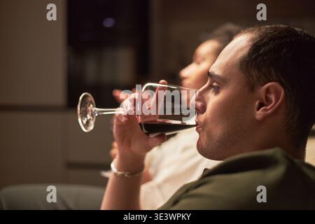 Kaukasischer junger erwachsener Mann trinkt Rotwein aus Glas, sitzt drinnen mit verschwommener junger erwachsener Frau im Hintergrund, entspannt zusammen in ungezwungener Atmosphäre Stockfoto