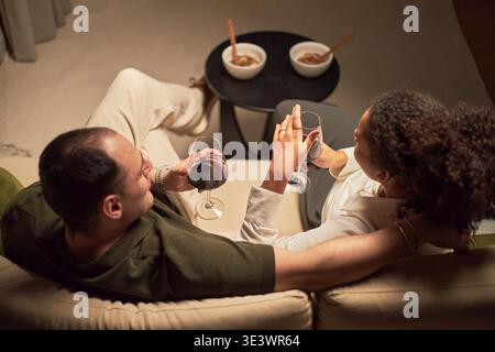 Kaukasischer Mann und Schwarze Frau sitzen dicht beieinander auf dem Sofa und halten Weingläser, entspannen und unterhalten, während sie mit zwei Schüsseln Essen auf einem kleinen Tisch in gemütlicher Inneneinrichtung essen Stockfoto