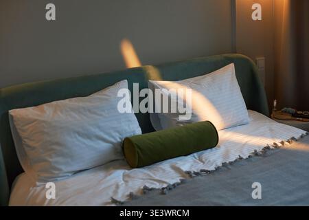 Doppelbett mit zwei großen Kissen und grünem zylindrischem Kissen gegen gepolstertes Kopfteil, Sonnenlicht schattenspendende Betten im sanft beleuchteten Schlafzimmer Stockfoto