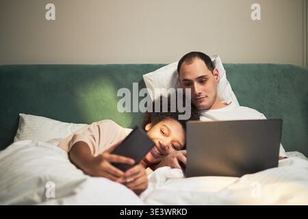 Junger Erwachsener kaukasischer Mann mit Laptop, während er im Bett neben dem jungen Erwachsenen liegt Schwarze Frau lächelt und hält ein Smartphone, beide entspannen zusammen unter der Decke im Schlafzimmer Stockfoto