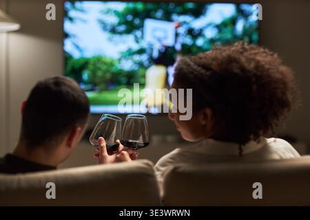 Junger Erwachsener kaukasischer Mann und junger Erwachsener Schwarze Frau sitzen auf Sofa und klirren Weingläser, während sie zusammen im Wohnzimmer Fernsehen, mit Blick auf die Rückseite der Kamera Stockfoto