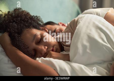Junge Erwachsene Schwarze Frau und junger Erwachsener kaukasischer Mann schlafen eng zusammen im Bett, beide ruhen friedlich mit Köpfen auf Kissen, intime Momente, die Paare umarmen Stockfoto