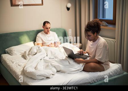 Junger Erwachsener kaukasischer Mann, der mit dem Smartphone auf dem Bett sitzt, während junge Erwachsene Schwarze Frau neben ihm sitzt und an einem Laptop arbeitet, beide konzentrierten sich auf Geräte in moderner Schlafzimmerumgebung Stockfoto