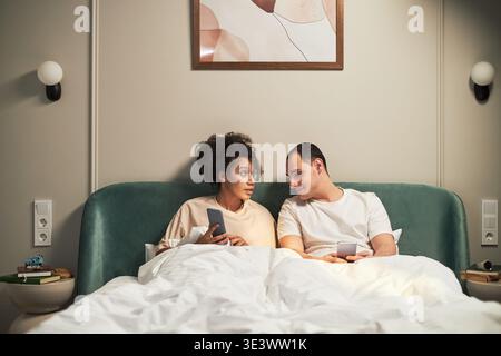 Junge Erwachsene Schwarze Frau und junger Erwachsener kaukasischer Mann liegen im Bett und halten Smartphones, schauen einander an und unterhalten sich, moderne Schlafzimmerumgebung sichtbar Stockfoto