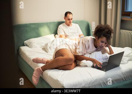 Junger Erwachsener kaukasischer Mann, der mit dem Smartphone auf dem Bett sitzt, während die junge Erwachsene Schwarze Frau neben ihm liegt und an einem Laptop arbeitet, beide konzentrierten sich auf Geräte in der modernen Schlafzimmerumgebung Stockfoto