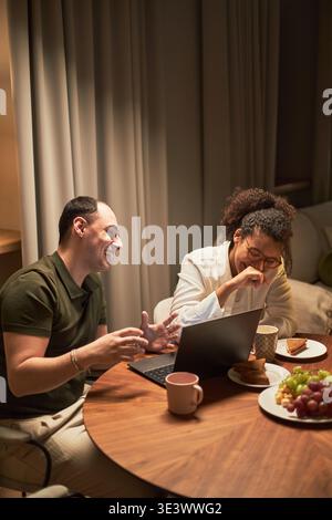 Vertikal des jungen Erwachsenen kaukasischen Mannes und der jungen Erwachsenen Schwarzen Frau, die am runden Tisch sitzen, lachen und reden, während sie Laptop benutzen, Essen mit Obst und Gebäck teilen und freundliche Gespräche genießen Stockfoto