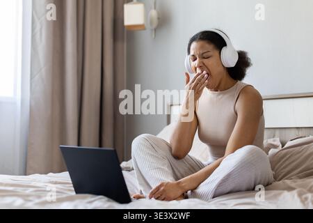 Schwarze Frau, die auf dem Bett sitzt und gähnt, während sie einen Laptop benutzt und Kopfhörer trägt Stockfoto