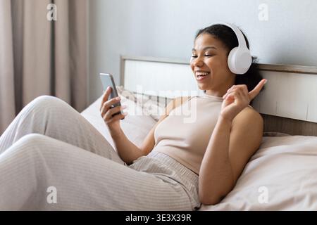 Schwarze Frau, die Kopfhörer trägt, Musik hört und das Smartphone im Bett benutzt Stockfoto