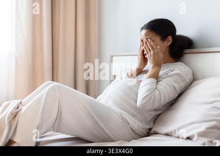 Schwangere schwarze Frau, die im Bett liegt und das Gesicht mit den Händen bedeckt, Stress ausdrückt Stockfoto