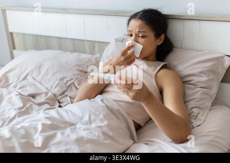 Junge schwarze Frau, die im Bett liegt und das Thermometer überprüft, während sie Gewebe hält Stockfoto
