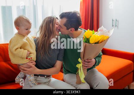 Alles Gute zum Muttertag. Ein Mann, der seine Frau küsst und zuhause ihren Blumenstrauß gibt. Frau, die ein Kleinkind hält. Liebevolle Eltern Stockfoto