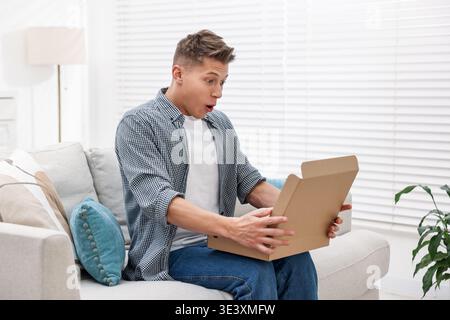 Ein überraschter Mann, der zu Hause das Paket auf dem Sofa öffnet Stockfoto