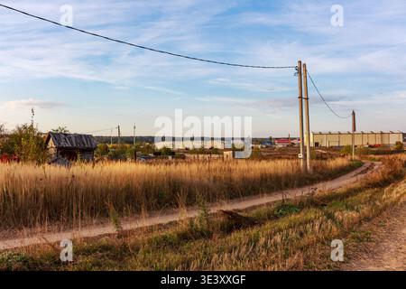 Alte, rustikale Hütte inmitten goldenem Gras und einer gewundenen Feldstraße unter einem riesigen Himmel. Stromleitungen werden an einen Pol angeschlossen, mit entfernten Industriegebäuden o Stockfoto