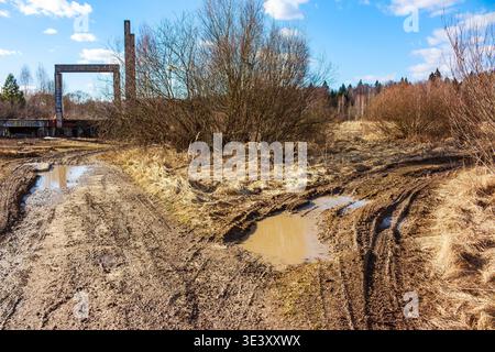 Knorriger Feldweg mit Pfützen durchquert ein karges Feld unter hellem Himmel und deutet auf anspruchsvolle Reisen in der Nähe vergessener Industriebauten hin Stockfoto