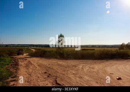 Ein staubiger weißer Van fährt über einen ländlichen Feldweg und rührt den Boden auf, während er in die malerische Landschaft fährt. Felder erstrecken sich unter einem hellblauen Himmel, Hi Stockfoto