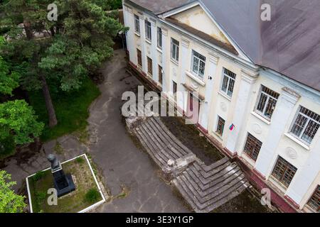 Die Fassade des alten Schulgebäudes und eine Denkmalbüste stehen im bewachsenen Innenhof. Abgenutzte Stufen führen zu der historischen Architektur, die von grünen Bäumen eingerahmt wird. Stockfoto