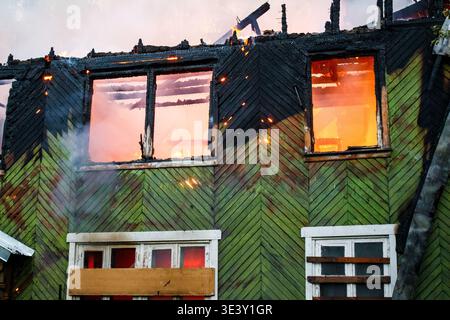 Intensives Feuer bricht durch ein verlassenes Holzhaus, feuriges Leuchten erfüllt die Fenster. Verkohlte Struktur, Rauch, Funken. Verheerende Flamme Stockfoto