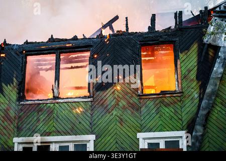 Inferno verzehrt ein altes Holzhaus, feuriges Licht, das durch die Fenster sichtbar ist, verkohlte Überreste des Daches vor einem rauchigen Himmel. Verheerende Flamme eines derartigen Stockfoto