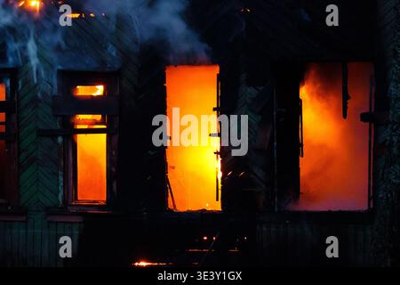 Feuriger Inferno-Eingeweide in einem alten, verfallenen Holzhaus. Helle orangene Flammen und Rauch schweben aus seinen dunklen Fenstern und Öffnungen, ein harter Ort des Notfalls Stockfoto