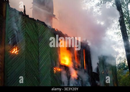 Ein hölzernes Gebäude, umgeben von einer Flamme. Intensive Flammen brechen aus den Fenstern und breiten sich über die Struktur aus und erzeugen dicken, welligen Rauch in den Himmel Stockfoto