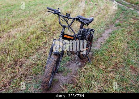 Das mit Schlamm bespritzte elektrische Fatbike liegt nach einer abenteuerlichen Offroad-Fahrt auf einem grasbewachsenen Feldweg und bietet rauen Outdoor-Spaß und anspruchsvollen Terr Stockfoto